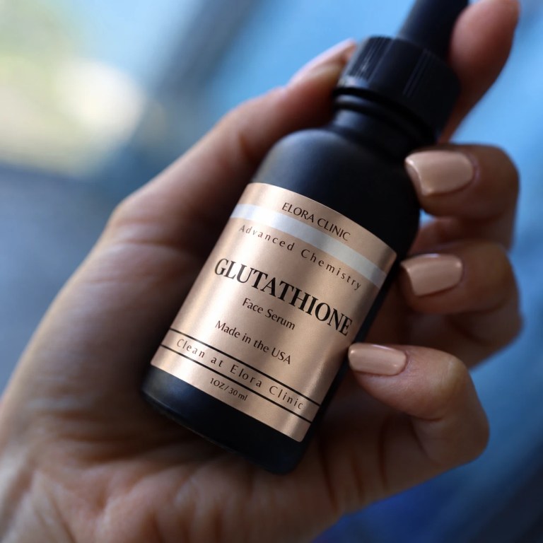 A hand holding a dropper bottle of Elora Clinic Glutathione Serum.