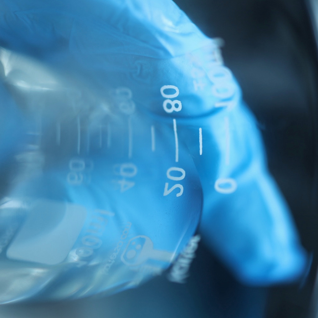 Close-up of glass beaker graduation marks with a blue-gloved hand behind the glass.