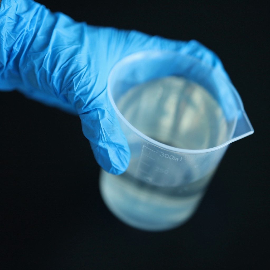 A blue-gloved hand holds a translucent measuring beaker containing clear liquid.