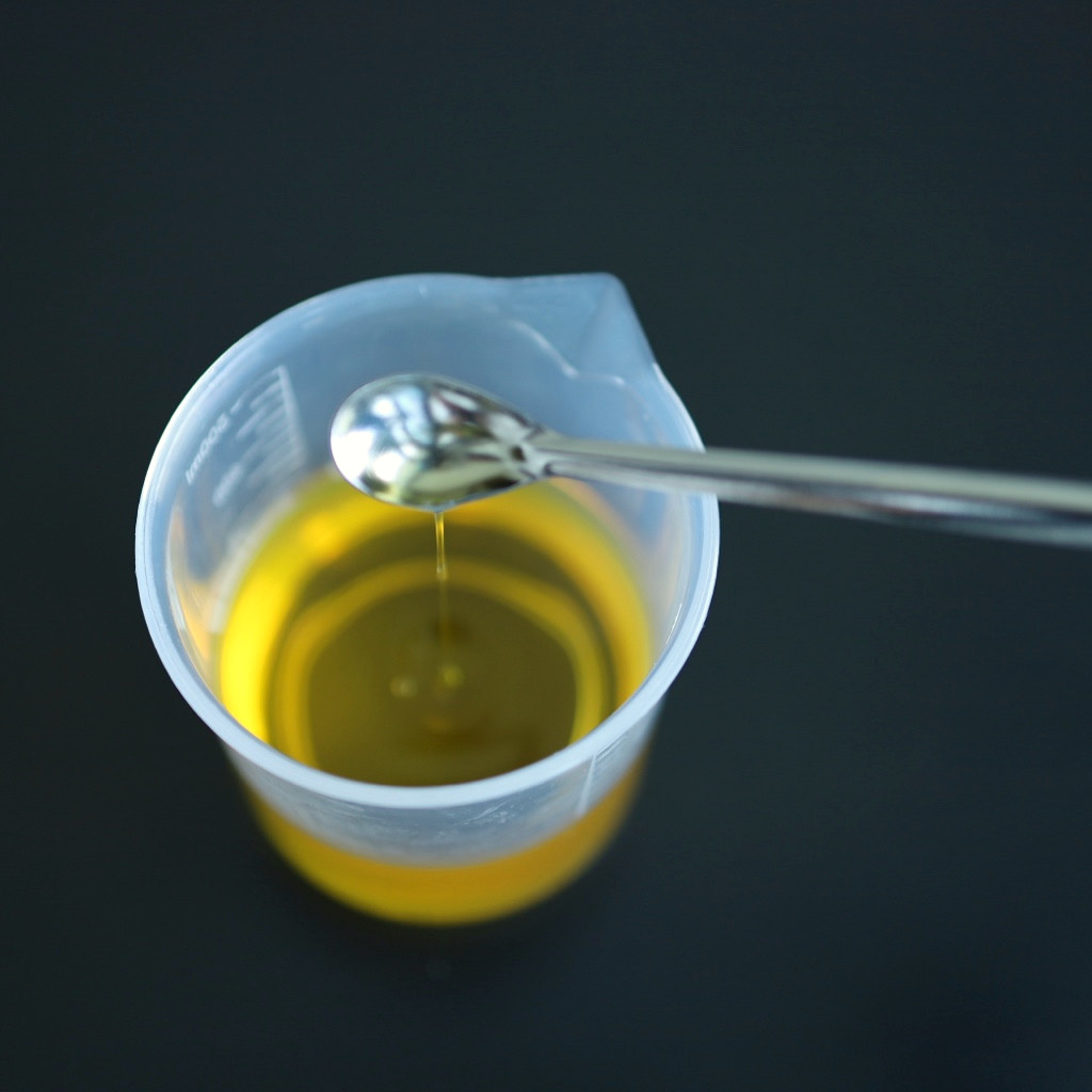 A thin strand of golden liquid drips from a metal spoon into a translucent measuring beaker.