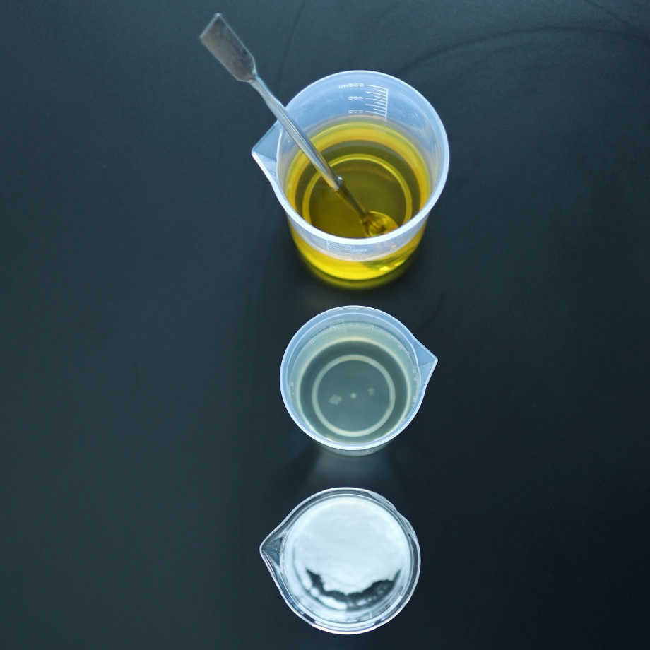 Three beakers arranged vertically contain golden liquid, clear liquid and white powder on a dark lab surface.
