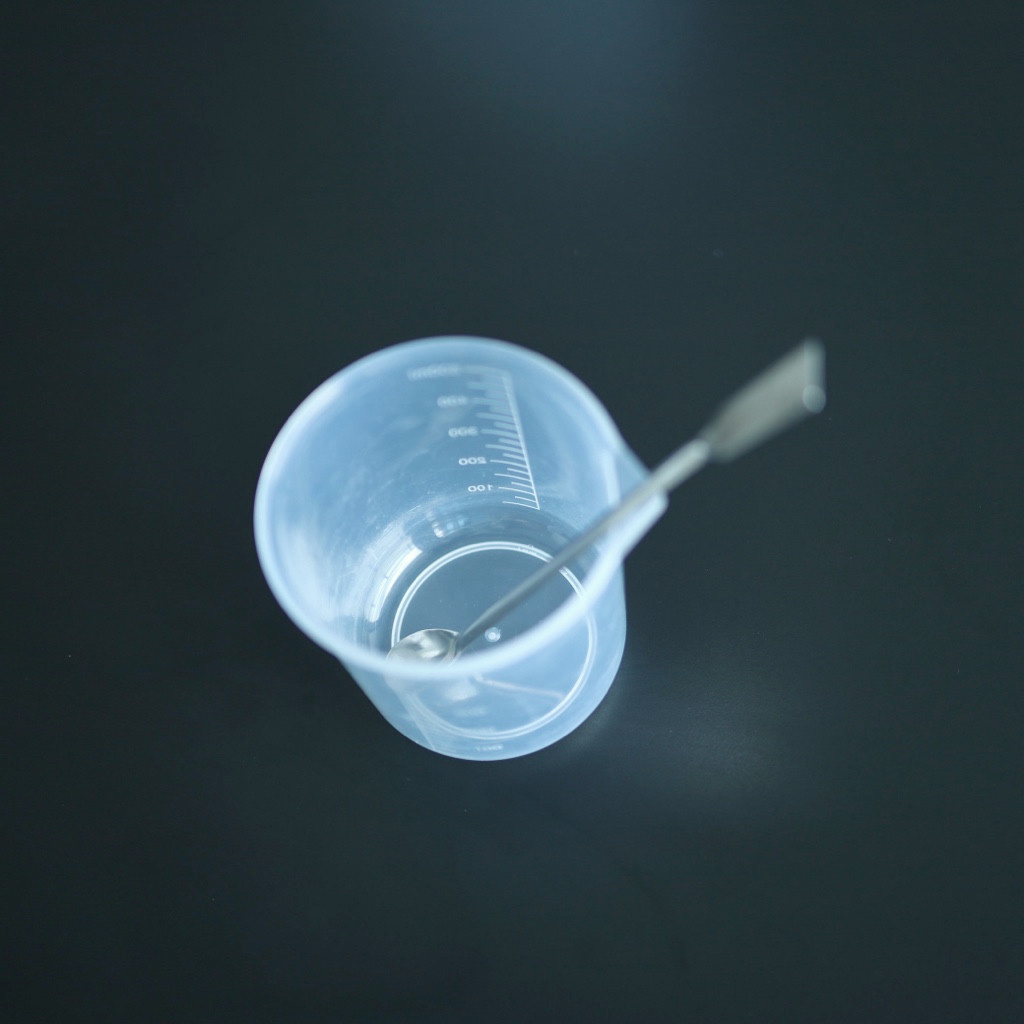 A translucent measuring beaker with a metal spatula rests on a dark lab work surface.
