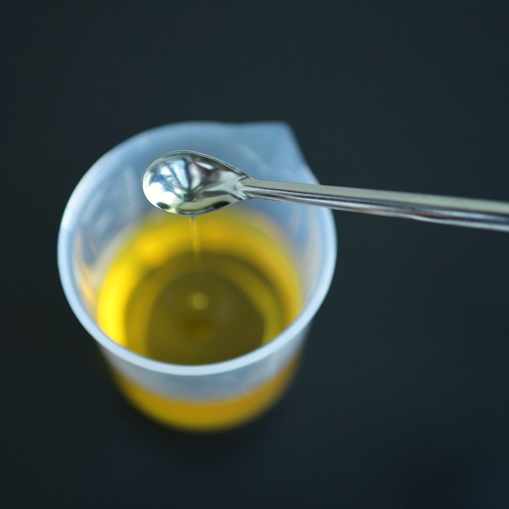 A metal spoon suspended over a beaker of golden liquid with a thin drip beneath it.